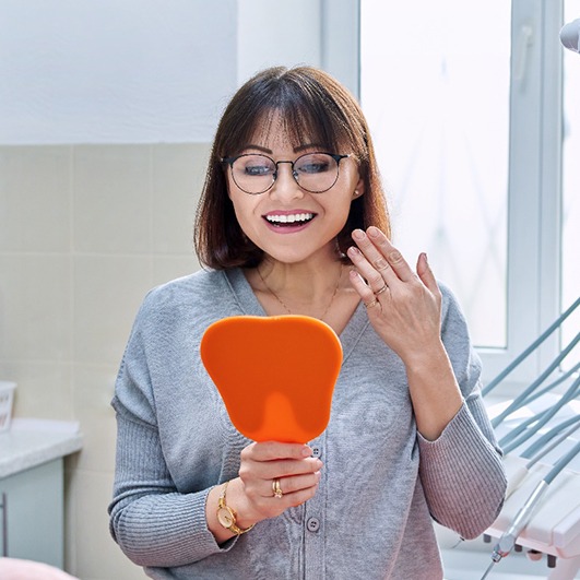 A smiling woman looking at her teeth in a hand mirror